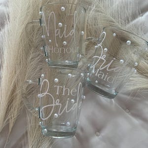 May include: Three clear glass mugs with white pearl accents and white lettering. The mugs say "Maid of Honour", "The Bride", and "Maid of Honour".