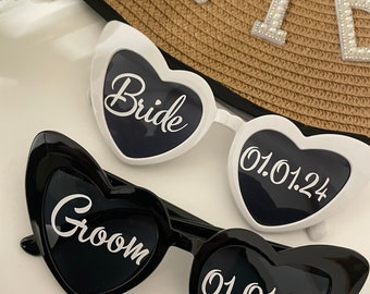 May include: A pair of heart-shaped sunglasses, one white with the word "Bride" and the date "01.01.24" and the other black with the word "Groom" and the date "01.01.24".