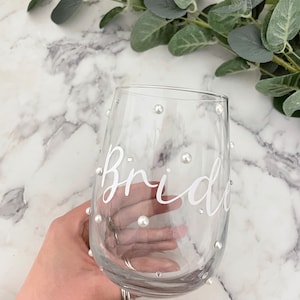 May include: A clear glass wine glass with the word "Bride" written on it in white lettering. The glass is decorated with small white pearl-like embellishments.