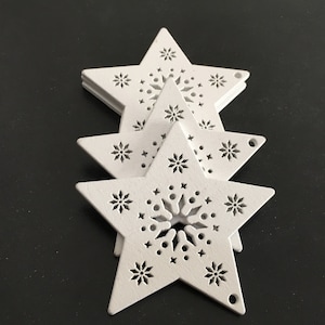 May include: Three white wooden star decorations with intricate cut-out designs. The stars are layered on top of each other, with the largest star at the bottom.
