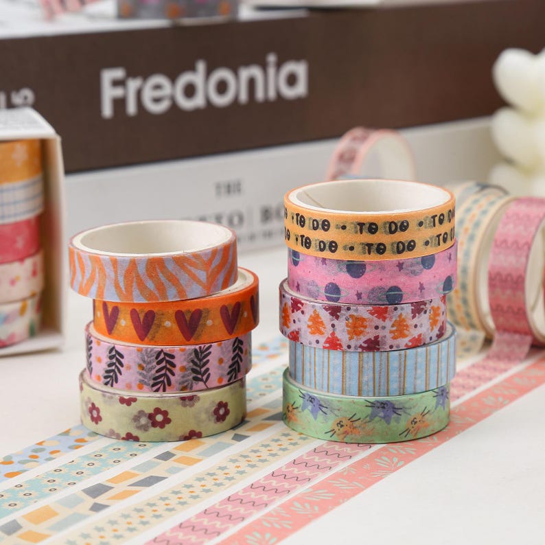 May include: A stack of colorful washi tapes with various patterns, including hearts, leaves, stars, and geometric shapes. The tapes are arranged in a circle on a white surface. The text "Fredonia" is visible on a box in the background.