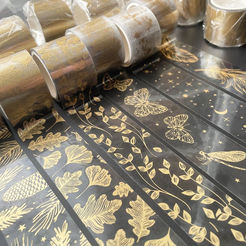 May include: Assortment of gold washi tape rolls with various black and gold patterned designs, including leaves, butterflies, stars, and a dragonfly. The tape is transparent and has a glossy finish.