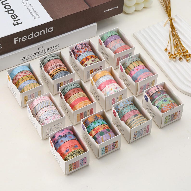 May include: A set of 12 colorful washi tape rolls in various patterns, each roll is packaged in a small white box with a clear window to view the tape. The boxes are arranged in a grid pattern on a white surface.