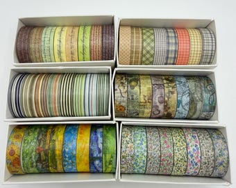 May include: A collection of six white cardboard boxes containing multiple rolls of decorative washi tape. The tapes feature various patterns including plaid, stripes, vintage script, floral prints, and artistic landscape designs in diverse colors.