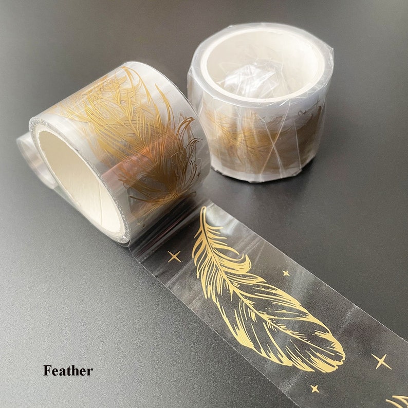 May include: Two rolls of clear washi tape with gold foil feather designs. The tape is rolled up and the design is visible on the outside of the roll. The tape is labeled "Feather".