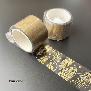 May include: Two rolls of gold washi tape with a pine cone and leaf design. The tape is clear with a gold pattern.