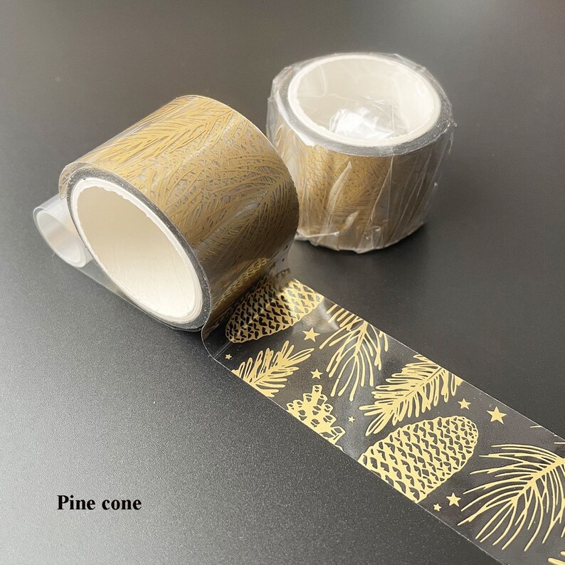 May include: Two rolls of gold washi tape with a pine cone and leaf design. The tape is clear with a gold pattern.