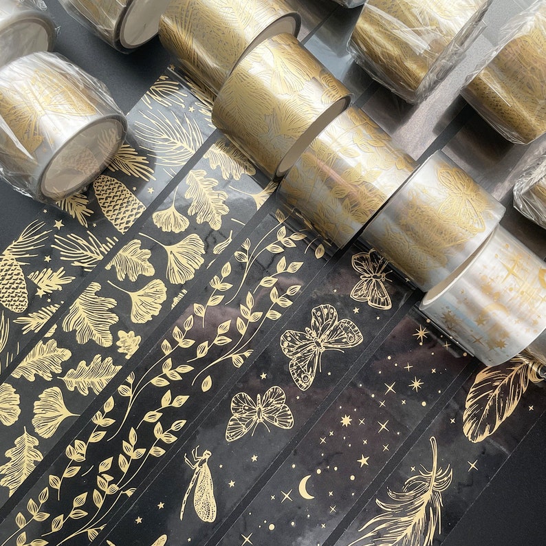 May include: Assortment of gold washi tape rolls with various patterns, including leaves, stars, feathers, and butterflies. The tape is on a black background.