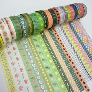 May include: A collection of decorative adhesive tape rolls arranged in a row, featuring various patterns including green leaves, flowers, geometric grids, stripes, and small crowns in shades of green, pink, orange, and blue on a white background.