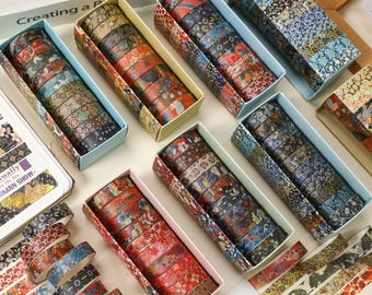 May include: A collection of decorative washi tape rolls in various colors and patterns, including floral and geometric designs. The tape is packaged in rectangular boxes with light blue and beige accents. The image also shows a book with the text "Creating a P" and "cually by the Sea HUMAN SHOW-".