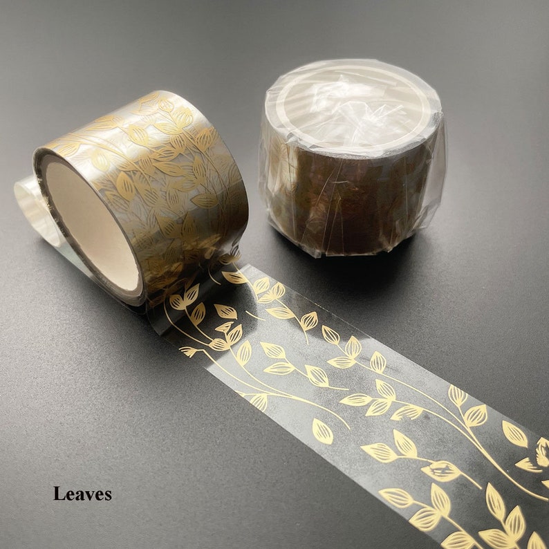 May include: Two rolls of washi tape with a gold leaf design on a black background. The tape is clear with gold leaf accents. The text "Leaves" is visible in the image.