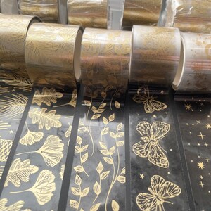 May include: Assortment of gold and black patterned washi tape rolls. The tape features designs of leaves, butterflies, and stars.