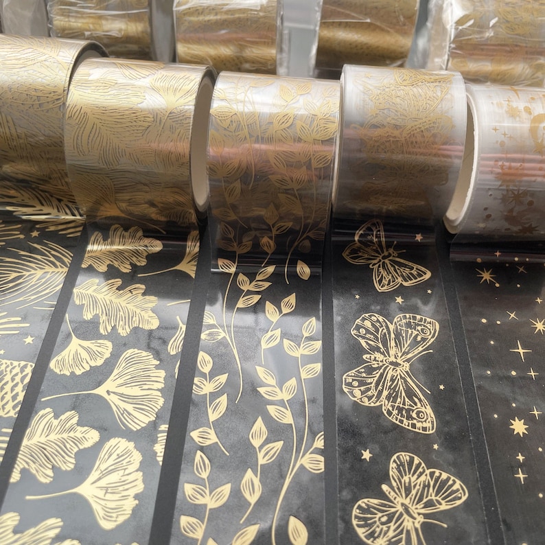 May include: Assortment of gold and black patterned washi tape rolls. The tape features designs of leaves, butterflies, and stars.