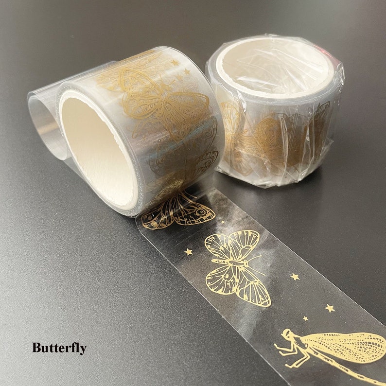 May include: Two rolls of clear tape with gold butterfly and dragonfly designs. The tape is on a gray surface.