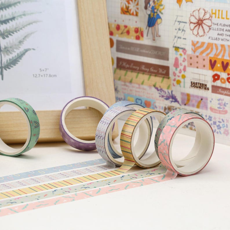 May include: Six rolls of patterned washi tape with various colors and designs. The tape is on a white surface with a wooden frame and a sheet of patterned paper in the background.