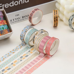 May include: A set of eight decorative washi tapes with various patterns, including geometric shapes, stars, and floral designs. The tapes are in shades of pink, blue, and beige.
