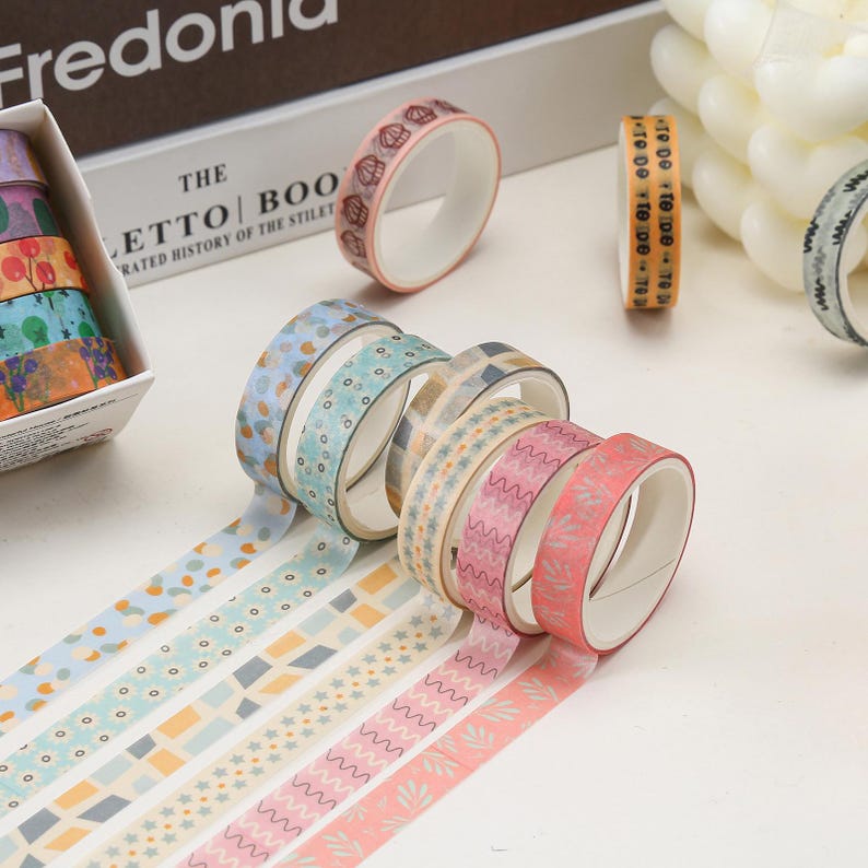 May include: A set of eight decorative washi tapes with various patterns, including geometric shapes, stars, and floral designs. The tapes are in shades of pink, blue, and beige.
