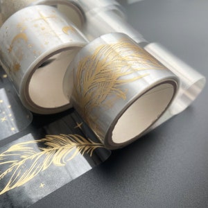 May include: A set of clear washi tape rolls with gold foil designs. The designs include feathers and stars.