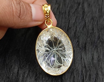 May include: A gold-colored pendant with an oval, clear crystal stone. The crystal has an intricate, etched design. The pendant is suspended from a gold-colored bail. The background is a dark, textured surface.