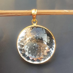 Op de afbeelding: Een hanger van helder kwartskristal met een gouden setting. Het kristal is gefacetteerd en heeft een ronde vorm.