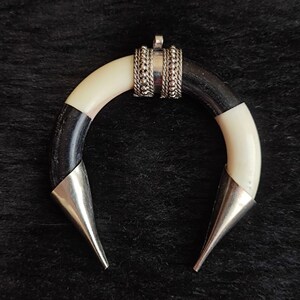 May include: A crescent-shaped pendant with a black and white horn design, accented with silver-colored metal. The pendant has a small loop at the top for a chain and two pointed ends. The design is simple yet striking.