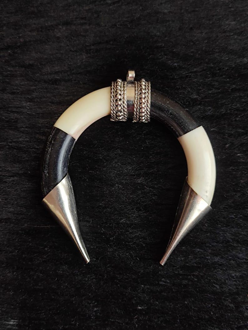 May include: A crescent-shaped pendant with a black and white horn design, accented with silver-colored metal. The pendant has a small loop at the top for a chain and two pointed ends. The design is simple yet striking.