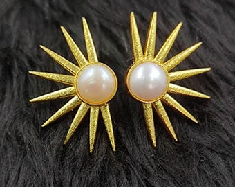 Peut inclure: Une paire de boucles d'oreilles dorées. Chaque boucle d'oreille présente une perle centrale, ronde et blanc cassé, entourée de pointes rayonnantes, ressemblant à un motif de soleil. Les boucles d'oreilles sont posées sur un fond sombre et texturé.