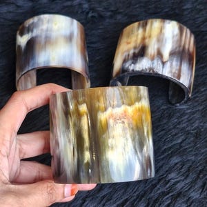 Puede incluir: Tres brazaletes anchos hechos de cuerno natural pulido, con una forma cilíndrica curva y lisa. El material muestra un patrón jaspeado con capas translúcidas en tonos crema, amarillo dorado, marrón oscuro y negro en toda la superficie.