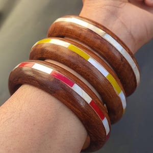 May include: Three wooden bangles with inlaid white, red, and yellow stripes. The bangles are stacked on a person's wrist.