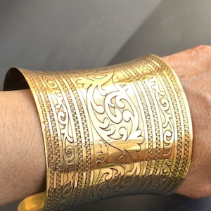以下が含まれることがあります： 複雑な花の模様が施された、幅広のゴールドトーンの金属製カフブレスレット。ブレスレットは真鍮製で、磨かれた仕上げになっています。