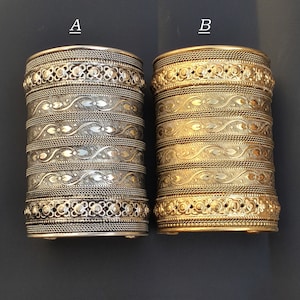 May include: Two silver and gold bracelets with intricate designs. The bracelets are stacked on top of each other, with the silver bracelet on top. The bracelets are both about 2 inches wide and have a variety of decorative elements, including floral patterns, geometric shapes, and beads.