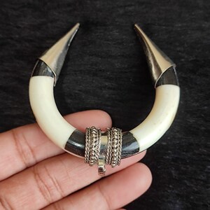 May include: A handcrafted, U-shaped pendant featuring a cream-colored, curved base with black accents. The ends are capped with silver-colored metal, and the center is adorned with silver-toned chain detailing and a small pendant.