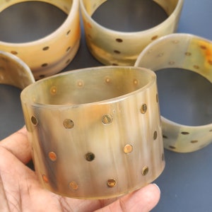 May include: Several wide, light-colored bangles with a speckled pattern. The bangles are a light tan color with darker brown and gold-colored spots. The bangles are stacked on top of each other, with one in the foreground.