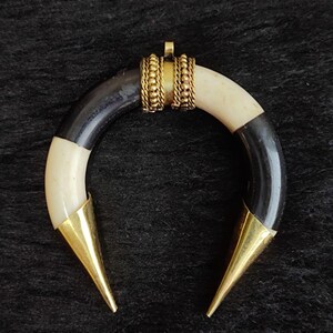 May include: A crescent-shaped pendant with alternating black and cream-colored sections. The pendant features gold-colored metal accents, including a decorative band and pointed tips. The jewelry piece is displayed against a dark, textured background.