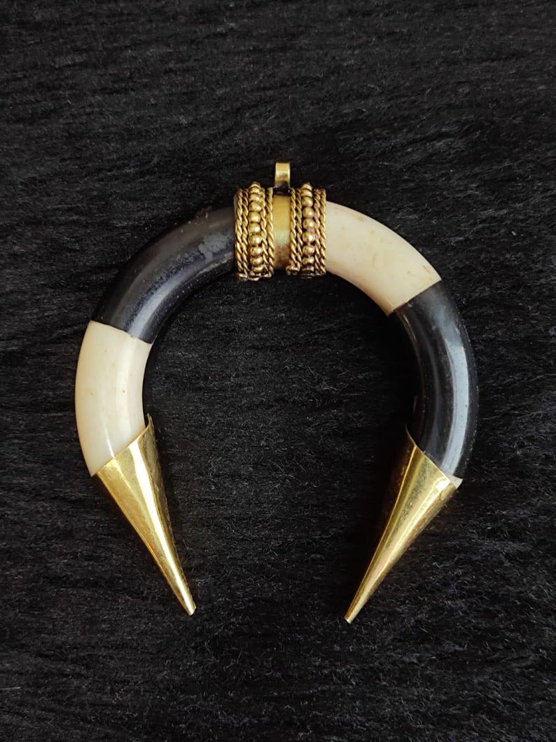 May include: A crescent-shaped pendant with alternating black and cream-colored sections. The pendant features gold-colored metal accents, including a decorative band and pointed tips. The jewelry piece is displayed against a dark, textured background.