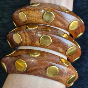 May include: Three wooden bangles with a rich brown finish and gold-colored metal studs. The bangles are stacked on top of each other, showcasing the polished wood and the decorative metal accents. The jewelry is displayed on a person's arm.
