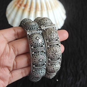 Puede incluir: Dos brazaletes articulados de tono plateado que presentan una serie de medallones circulares en relieve con intrincados patrones geométricos y de filigrana. La superficie metálica tiene un acabado oxidado oscuro que resalta las texturas.