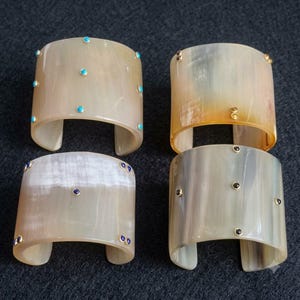 Può includere: Quattro bracciali a fascia larga realizzati in materiale di corno lucido, traslucido e di colore chiaro. Ogni bracciale presenta piccole pietre rotonde incastonate in metallo di vari colori, tra cui turchese, blu scuro, ambra e nero, disposte a motivo.
