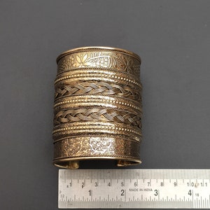 Op de afbeelding: Een gouden metalen armband met een ingewikkeld geweven ontwerp en bloemmotieven. De armband heeft een diameter van ongeveer 5 cm.