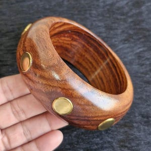 May include: A polished wooden bangle bracelet with a rich brown grain pattern. The bracelet is adorned with several small, round, gold-colored metal accents. The bracelet is approximately 3 inches in diameter.
