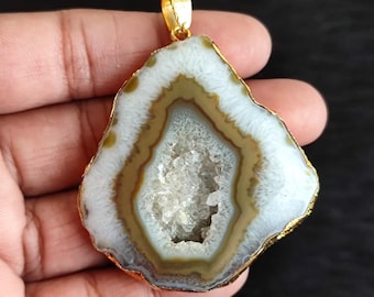 May include: A natural agate pendant with a gold-colored metal trim and bail. The stone features a white, brown, and clear crystal center. The pendant is irregularly shaped and is held in a hand against a dark background.