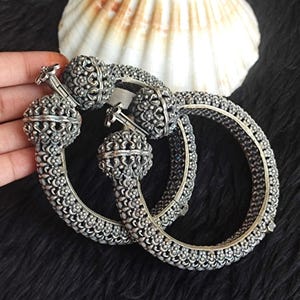 Könnte beinhalten: Ein Paar Armreifen aus silberfarbenem Metall mit einem komplexen, erhabenen Blumen-Filigranmuster auf dem gesamten Band. Jeder Armreif ist mit einem markanten, kugelförmigen Verschlussmechanismus versehen, der mit detaillierten Metallarbeiten verziert ist.