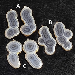 May include: Six gray and white agate pendants with gold-colored edges. Each pendant has a unique pattern of concentric circles and lines. The pendants are displayed on a dark, textured surface.