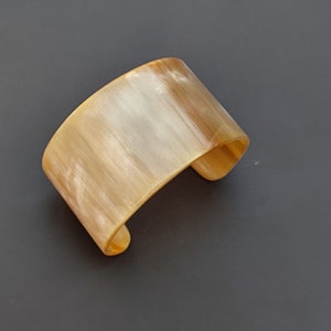 Op de afbeelding: Een brede, gebogen manchetarmband in tinten bruin en crème. De armband heeft een glad, gepolijst oppervlak met natuurlijke kleur- en patroonvariaties. Het open ontwerp maakt het gemakkelijk om te dragen.