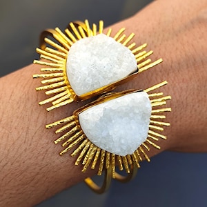 May include: Gold-tone bracelet featuring two large, irregular white stone accents. The stones are set in gold-colored frames, surrounded by radiating gold spikes. The bracelet is a bold, statement piece of jewelry.