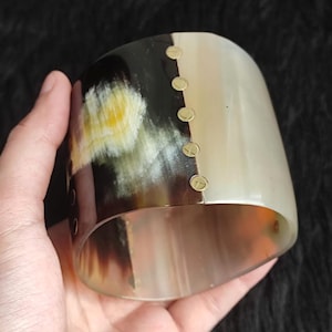 Op de afbeelding: Een brede armband van gepolijst hoorn met een tweekleurig ontwerp, waarbij een donkerbruin, zwart en geel gemarmerd gedeelte is verbonden met een doorschijnend crèmekleurig gedeelte door een verticale rij van vijf kleine, ronde, goudkleurige metalen klinknagels.