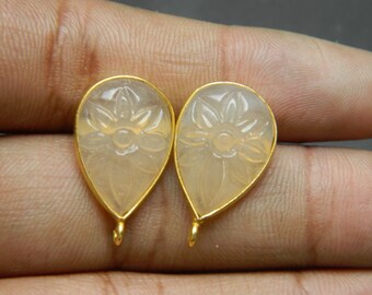 May include: Two teardrop-shaped earrings with a floral design carved into a light peach-colored stone. Each stone is set in a gold-colored metal frame with a small loop at the bottom for attachment. The earrings are displayed against a neutral background.