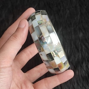 Pode incluir: Uma pulseira tipo bracelete com um padrão de mosaico de peças iridescentes de madrepérola e concha de abalone. As peças retangulares estão dispostas numa grelha, exibindo vários tons de branco, creme, cinzento e toques subtis de verde e azul.
