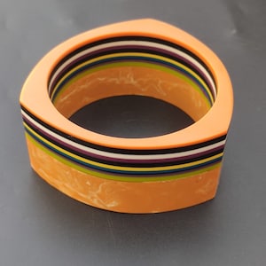 Puede incluir: Un brazalete de rayas multicolores con un acabado naranja marmoleado. El brazalete está hecho de plástico y tiene una forma geométrica.