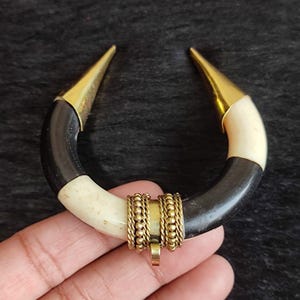 May include: A handcrafted, U-shaped pendant necklace. The pendant features a curved design with black and cream sections, accented with gold-colored metal tips and a central decorative element. The necklace is designed to be worn as a statement piece.